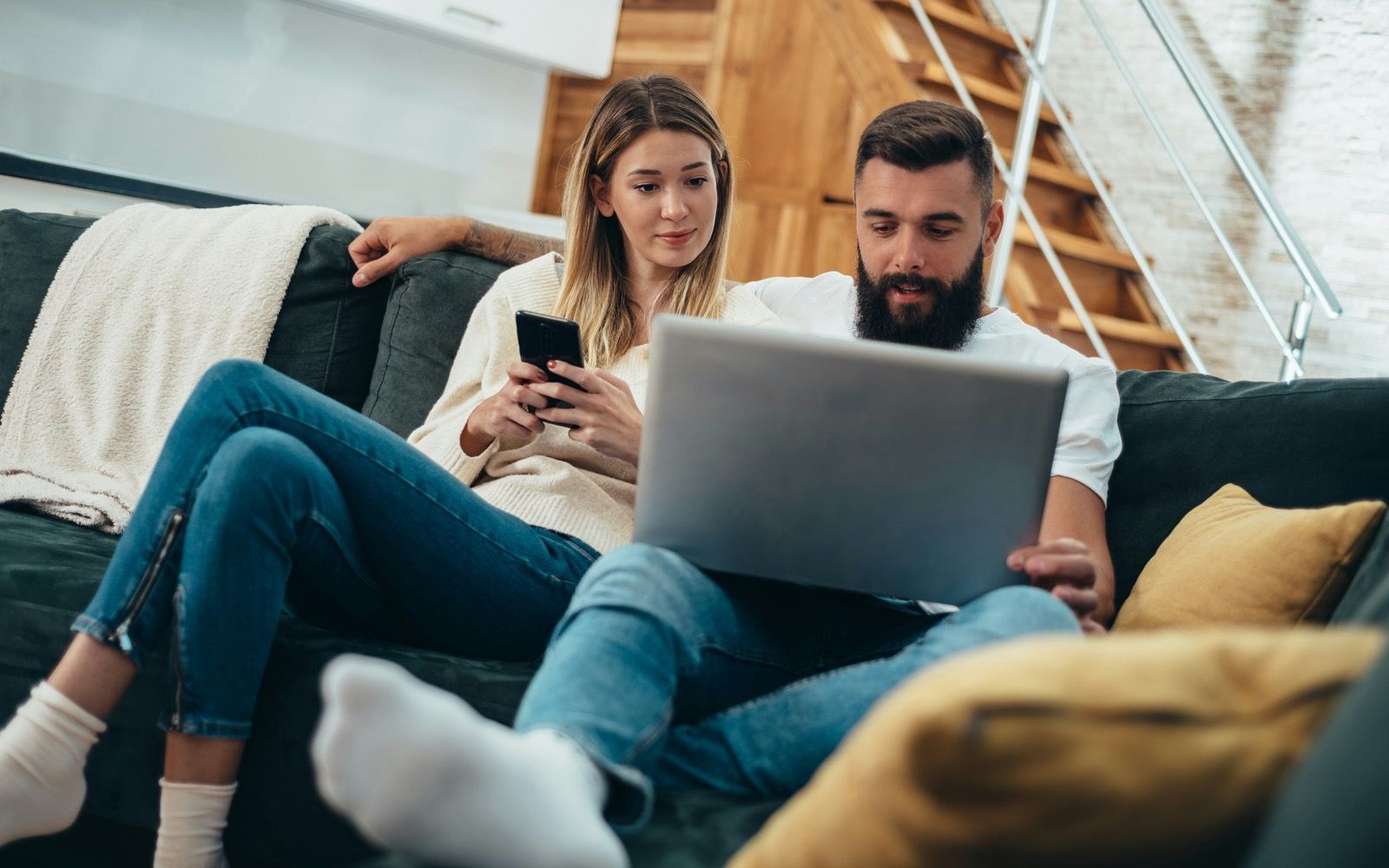 A couple relaxing on their couch at home, completing a quick health intake on a phone and laptop